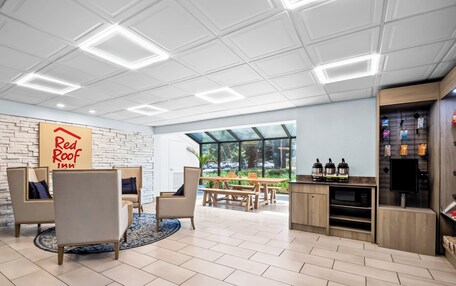 A modern lounge area with comfortable seating, a Red Roof Inn sign, a kitchen area with appliances, and a view of the outdoors.
