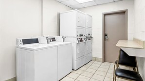 A laundry room with a washing machine, dryers, and a small table with chairs. - Red Roof Inn Myrtle Beach Hotel - Market Common (Myrtle Beach)