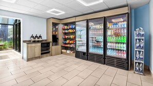 A modern kitchenette with a microwave, kettle, and kettle stand, a snack bar with chips and snacks, and a refrigerator stocked with beverages and water. - Red Roof Inn Myrtle Beach Hotel - Market Common (Myrtle Beach)