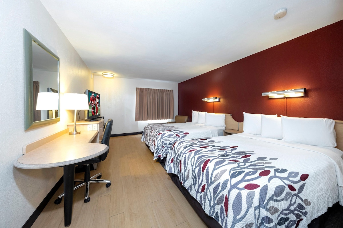 A hotel room with a desk, two beds, a mirror, and a red accent wall.