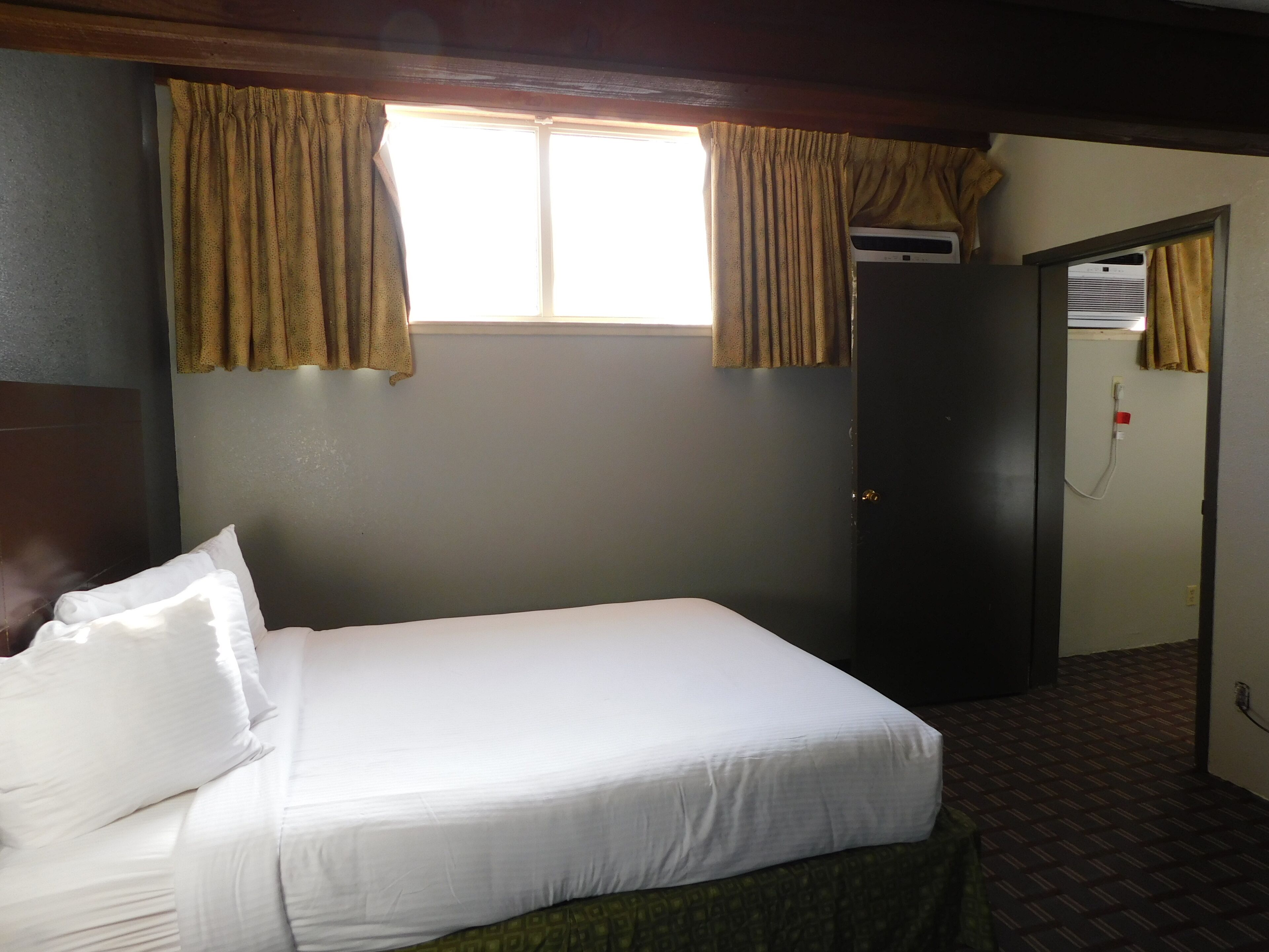Een hotelkamer met een bed, een raam met gordijnen, een airconditioningsunit en een deur.