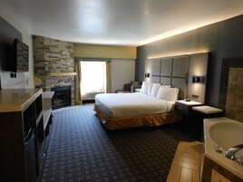 A hotel room with a large bed, a flat-screen TV, a fireplace, a desk, and a bathroom with a bathtub and sink.