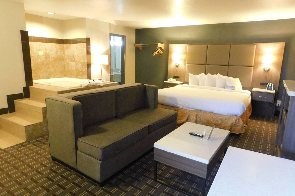 A hotel room with a bed, a sofa, a coffee table, a desk, a lamp, a mirror, and a bathtub.