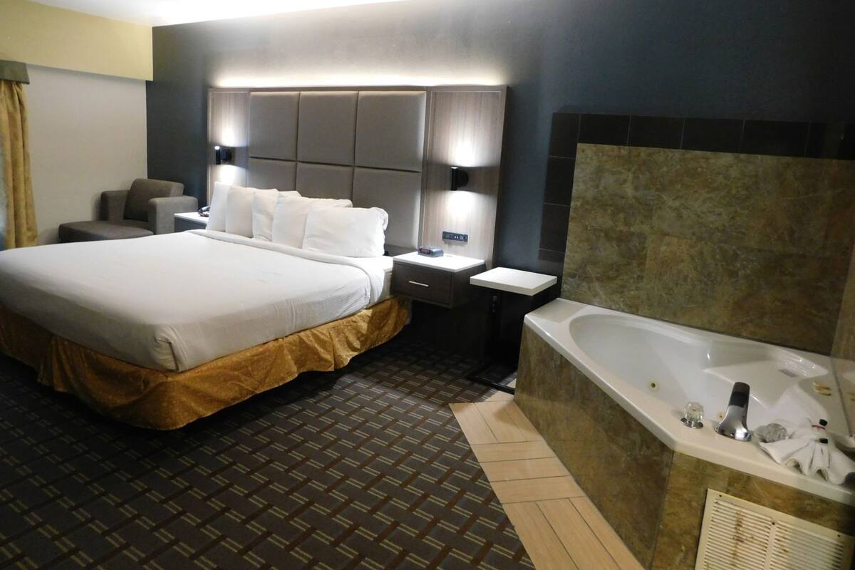 A hotel room with a large bed, bedside tables, a bathroom with a bathtub, and a headboard with built-in lighting.