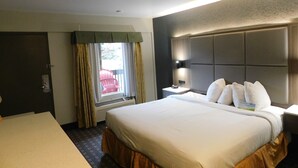 Habitación de hotel con una cama grande, una mesita de noche con lámpara, una ventana con cortinas y una puerta.