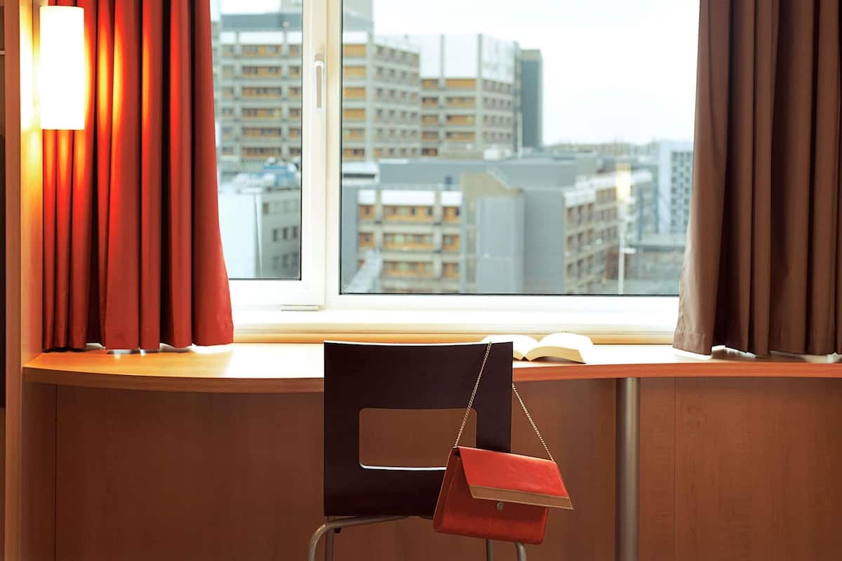 A hotel room with a view of city buildings, a desk with a chair, and a red handbag hanging on the chair.