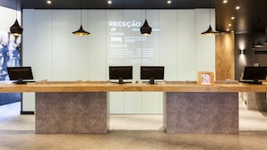 A reception desk with three monitors, a wooden counter, and a wall with service information. - ibis Lisboa José Malhoa (Lisbon)