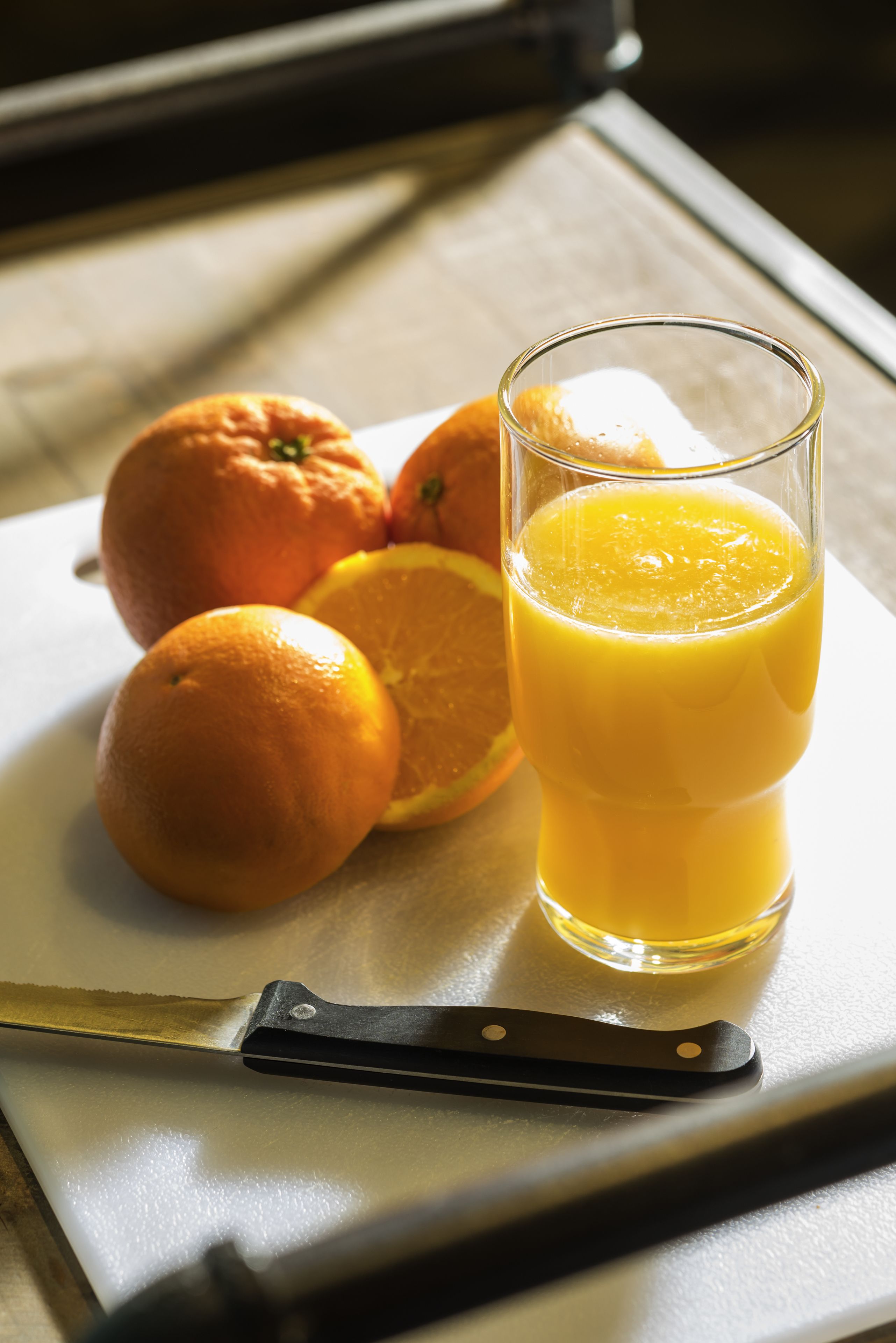 Un vaso de jugo de naranja, un cuchillo y naranjas sobre una tabla de cortar.