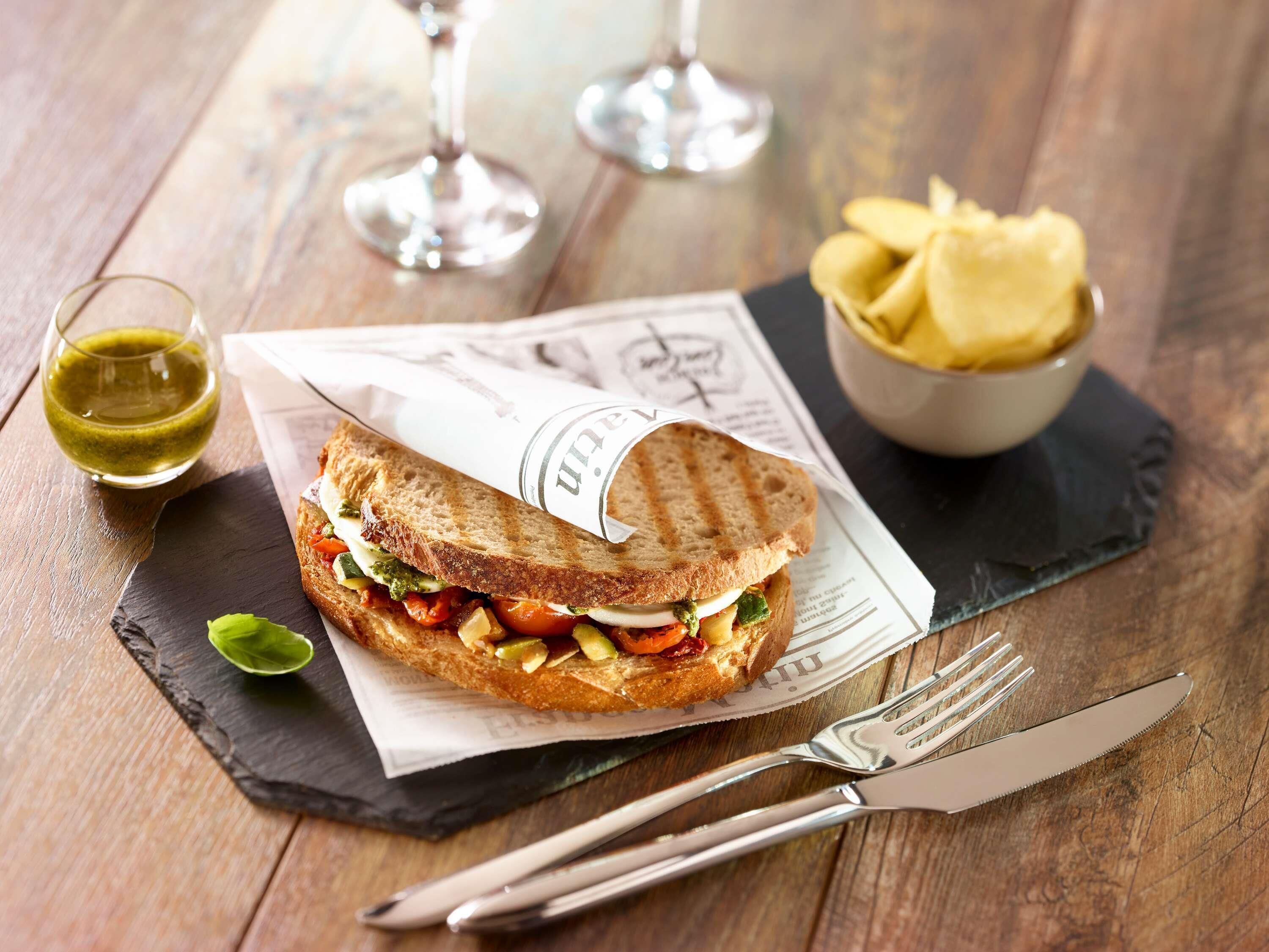 Ein Sandwich auf einer Schieferplatte, daneben eine Gabel und ein Messer, ein Glas mit grüner Soße und eine Schüssel Chips.