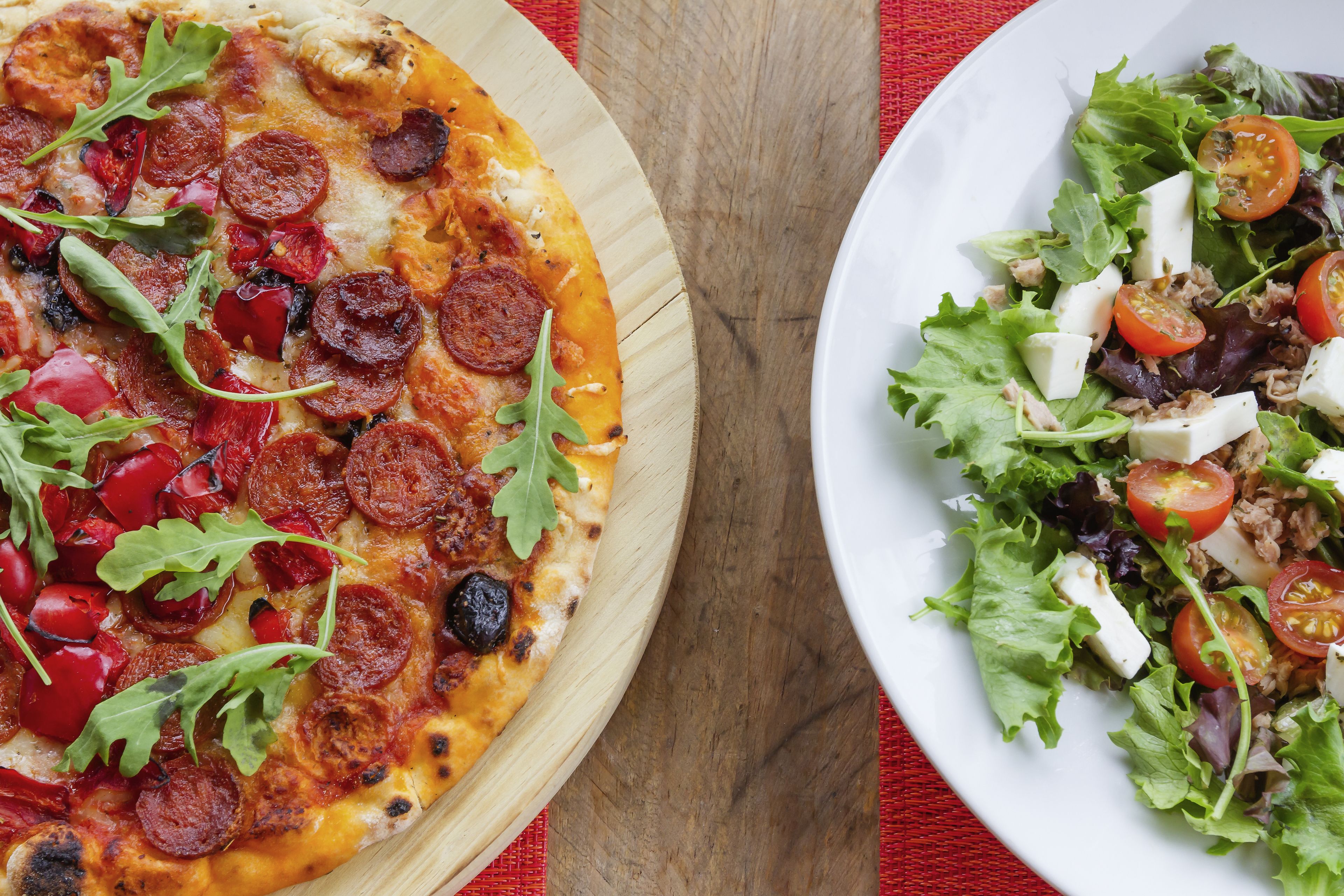 A pizza with pepperoni and greens on a wooden board, and a salad with greens and tomatoes on a white plate.