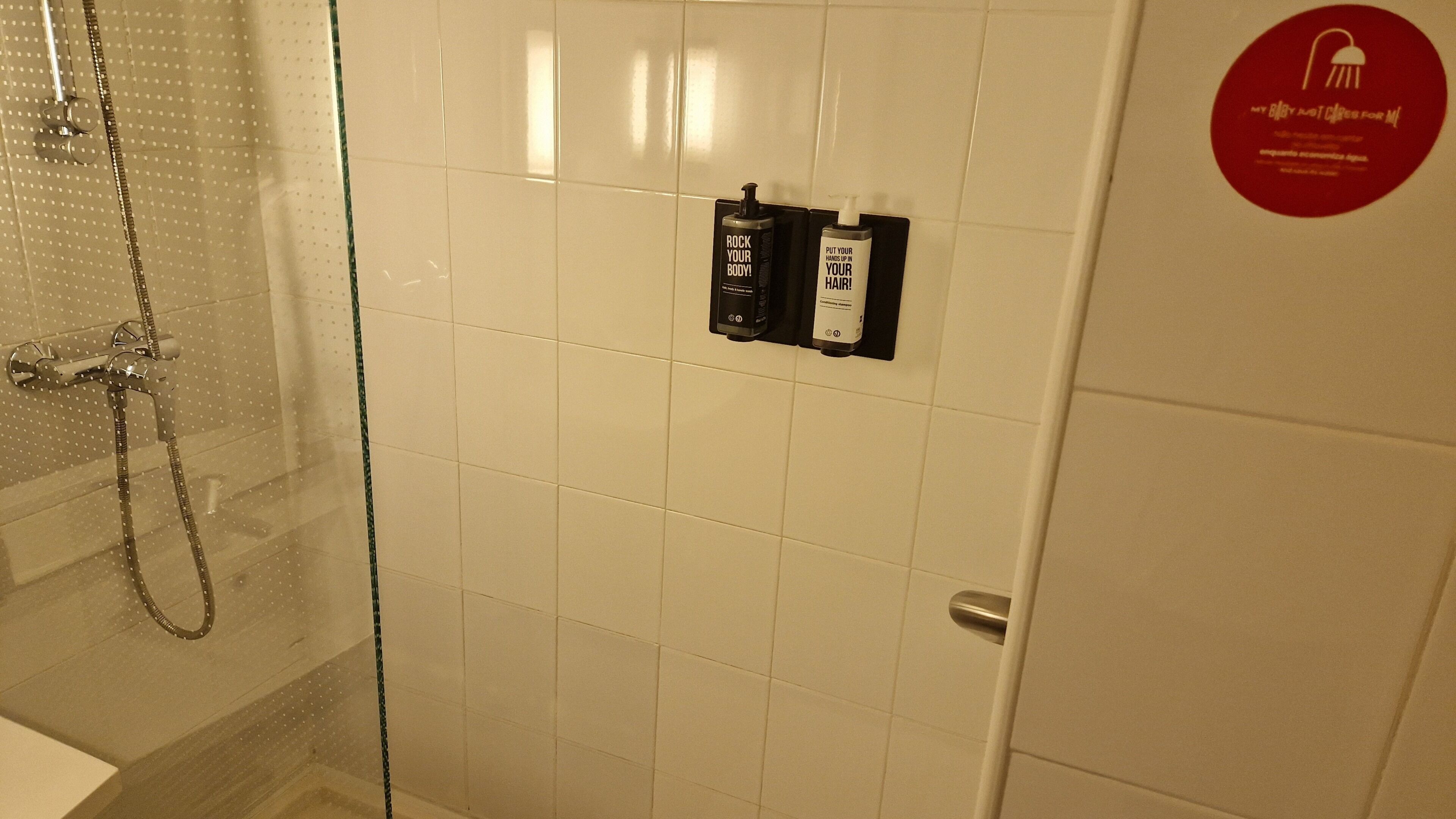 A bathroom with a glass shower enclosure, a white tiled wall, and a red circular sticker on the door.