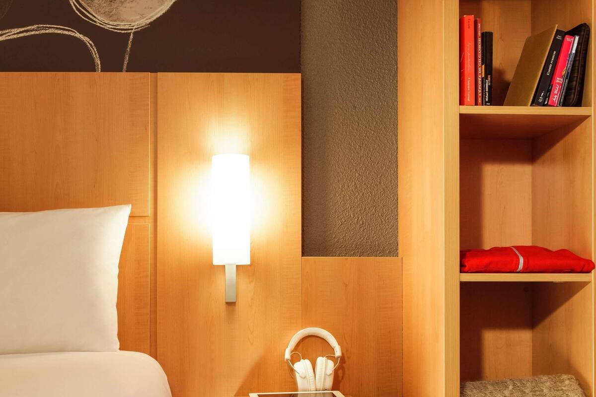 Une chambre d’hôtel avec un lit, une table de chevet sur laquelle se trouvent une tablette et des écouteurs, une lampe fixée au mur et une bibliothèque contenant des livres et une serviette rouge.