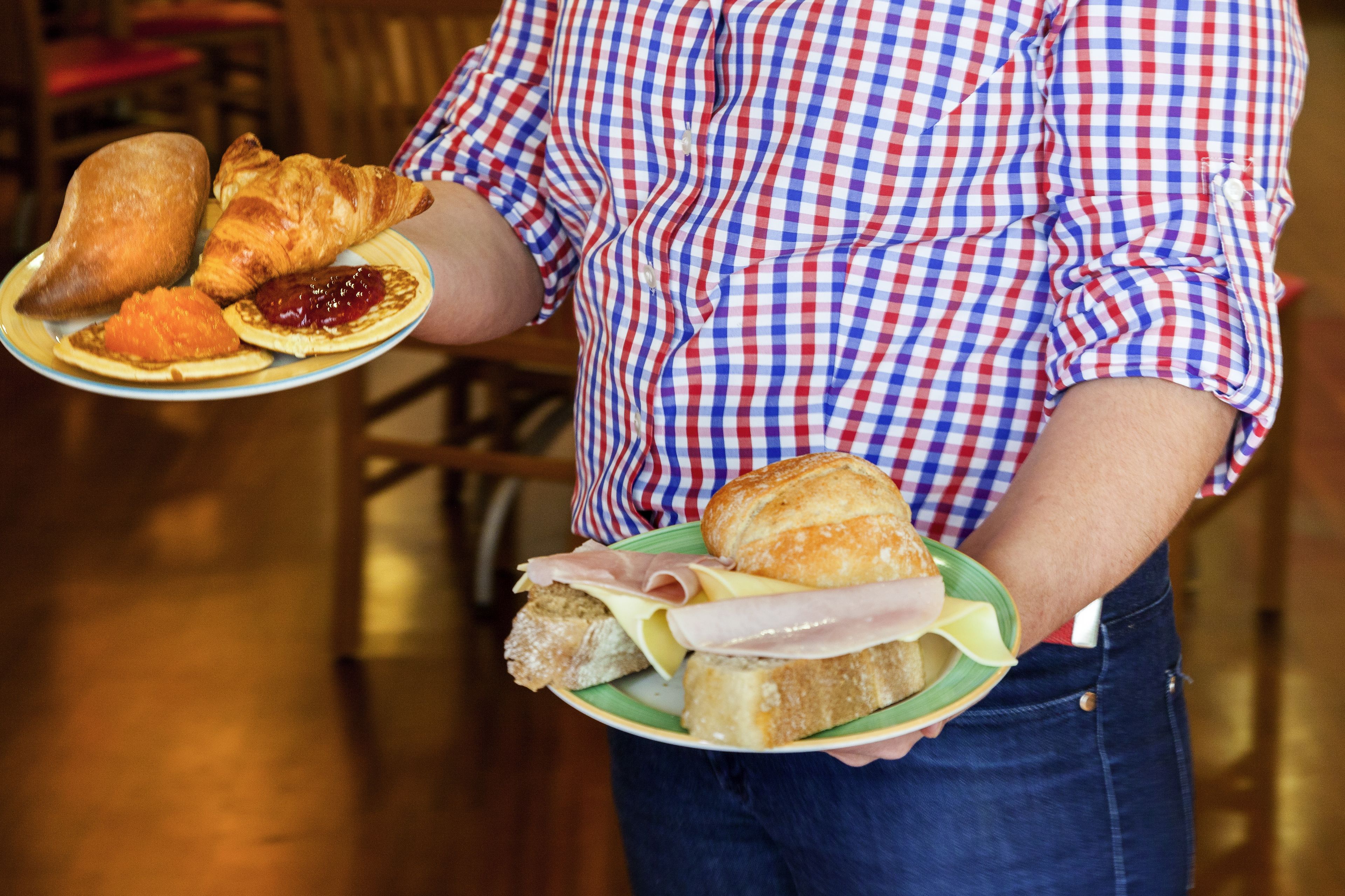 Una persona sosteniendo un plato con croissants, un sándwich de jamón y un pastel.