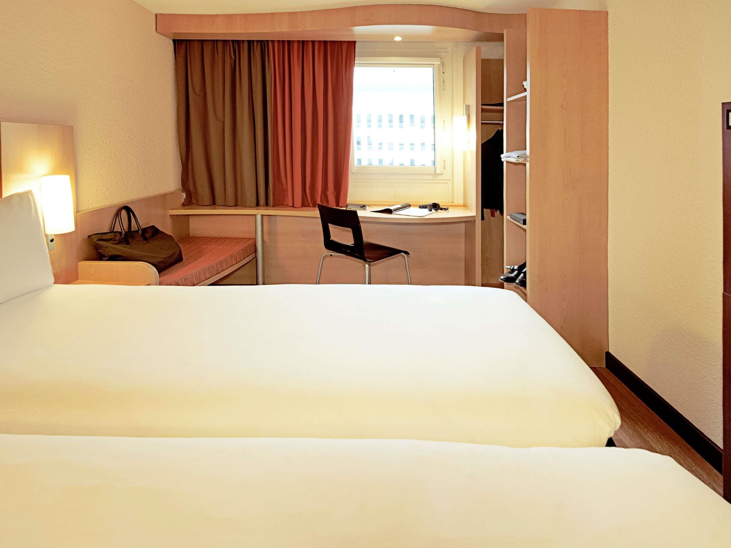 Une chambre d’hôtel avec un grand lit, un bureau, une chaise, une lampe, un porte-manteau et une fenêtre avec des rideaux.