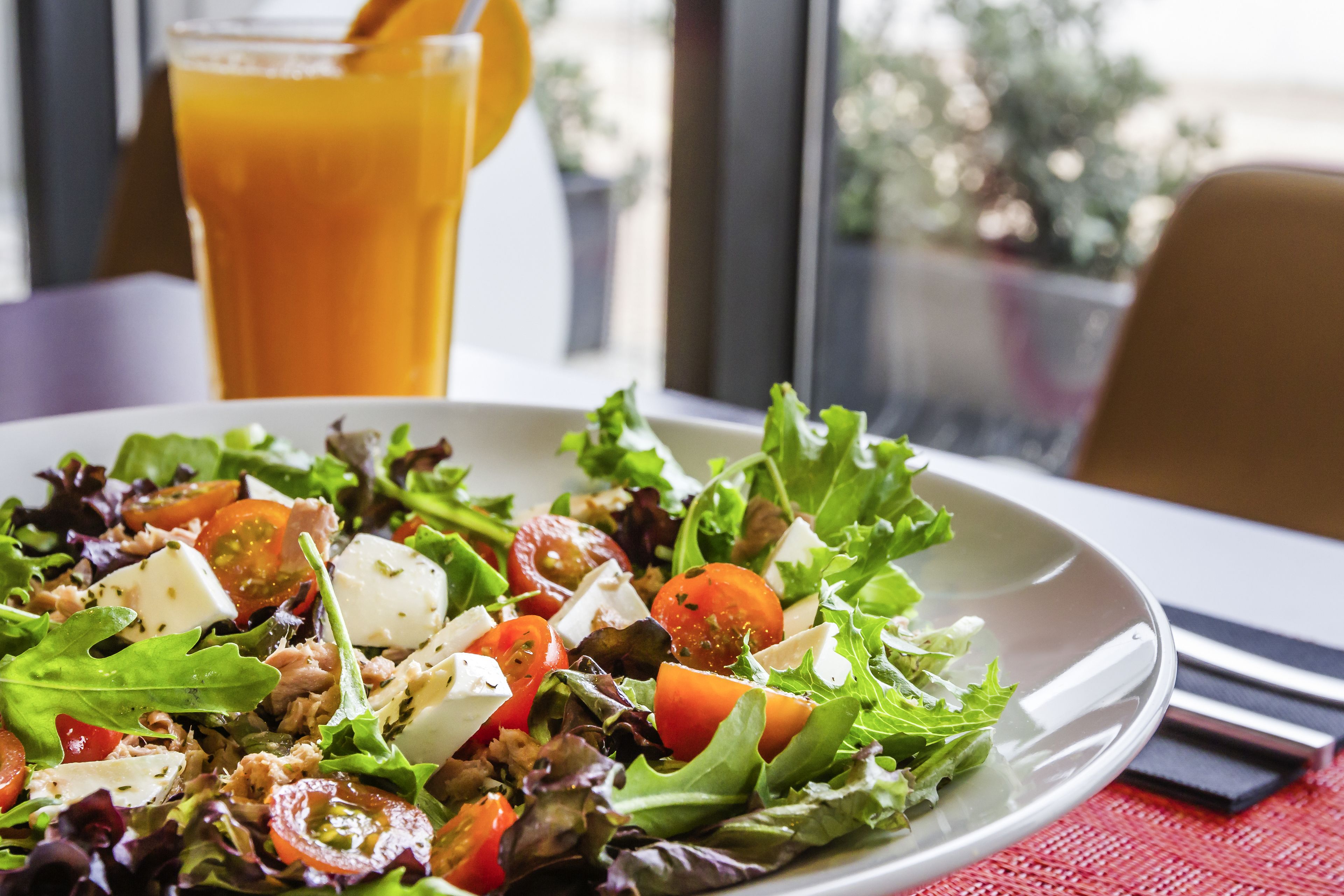 Uma salada com tomates, queijo e folhas verdes, e um copo de sumo de laranja numa mesa com vista para a janela.