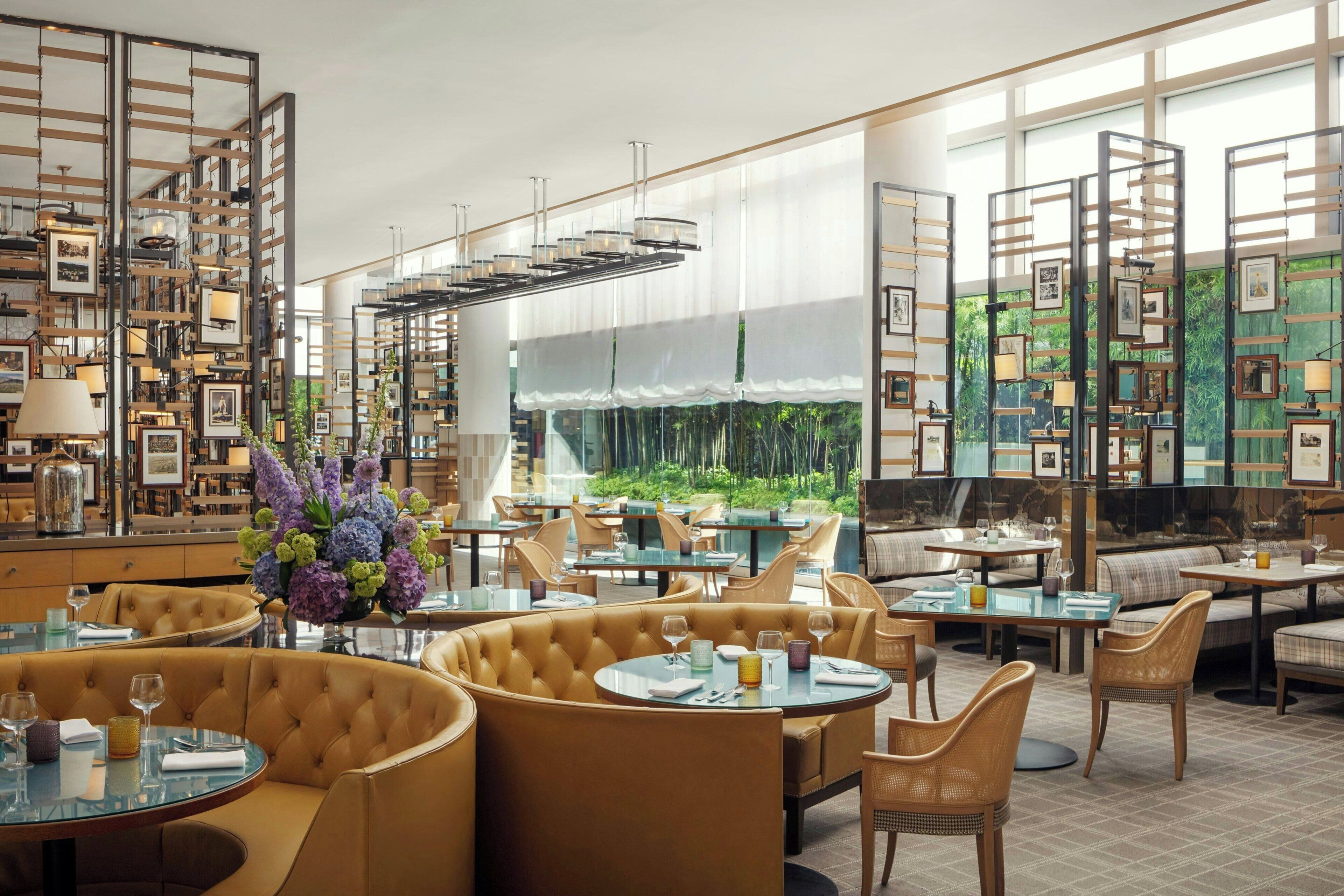 A modern restaurant with round tables, cushioned seating, and a view of greenery through large windows.