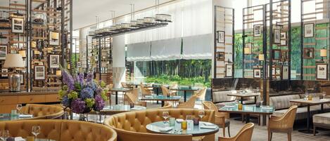 A modern restaurant with round tables, cushioned seating, and a view of greenery through large windows.