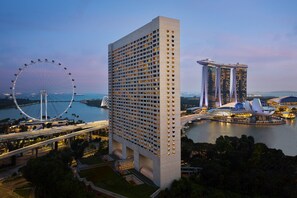A high-rise hotel with a modern design, located near a waterfront with a Ferris wheel and a distinctive building with a unique roof structure.