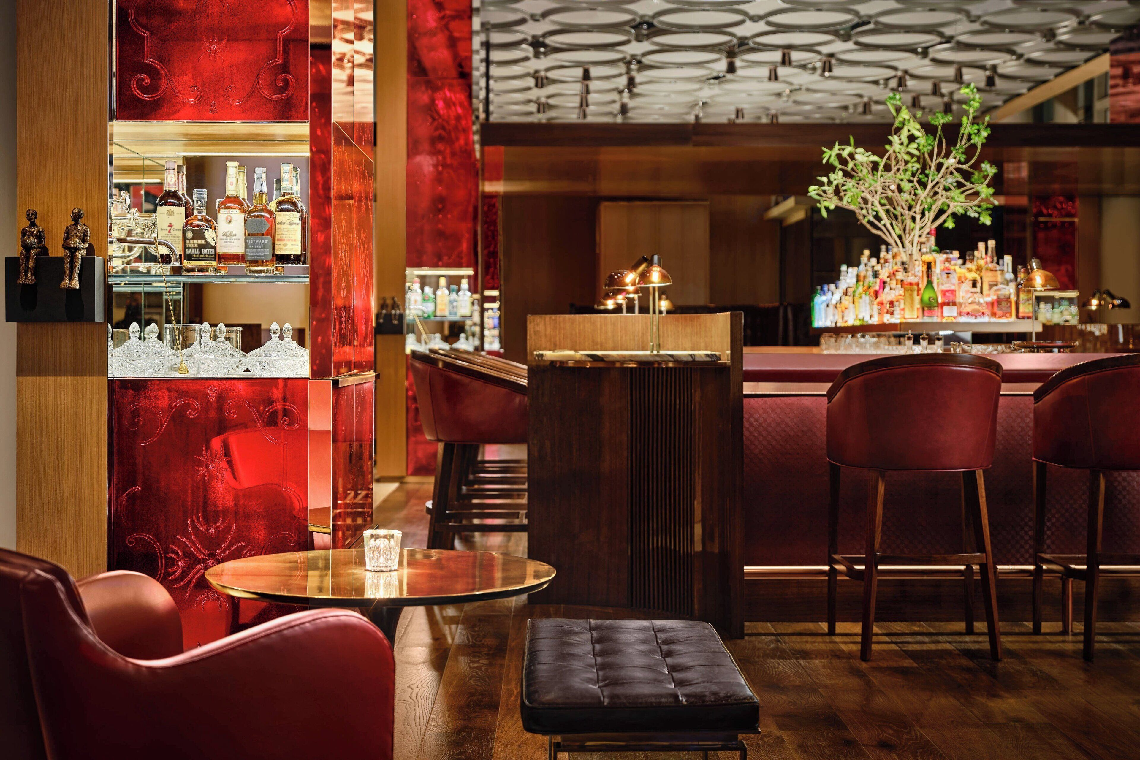 Una zona de bar con mostrador de madera, sillas tapizadas en rojo, una mesa redonda con un vaso y una planta decorativa.