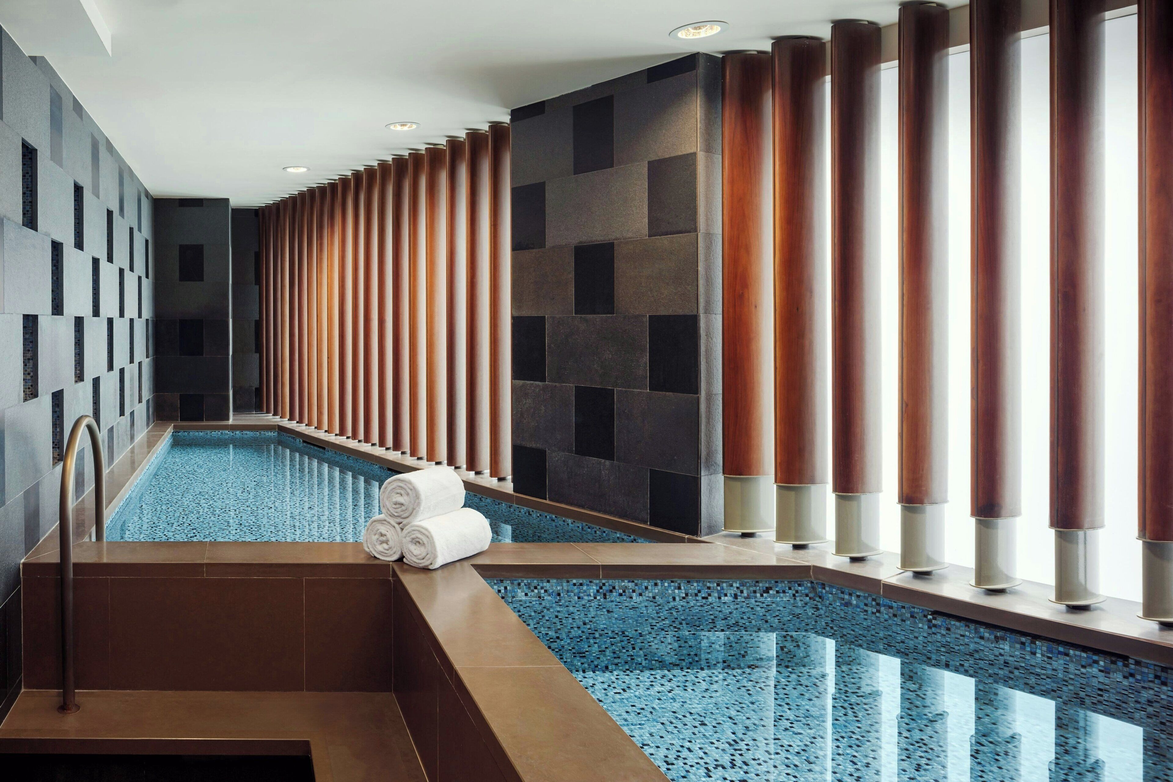 A swimming pool with a tiled floor, a set of wooden columns, and a towel rack with towels.