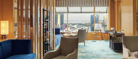 Un hall de hotel con un sofá azul, un sillón beige, una mesita de centro y vistas a un paisaje urbano a través de amplias ventanas.
