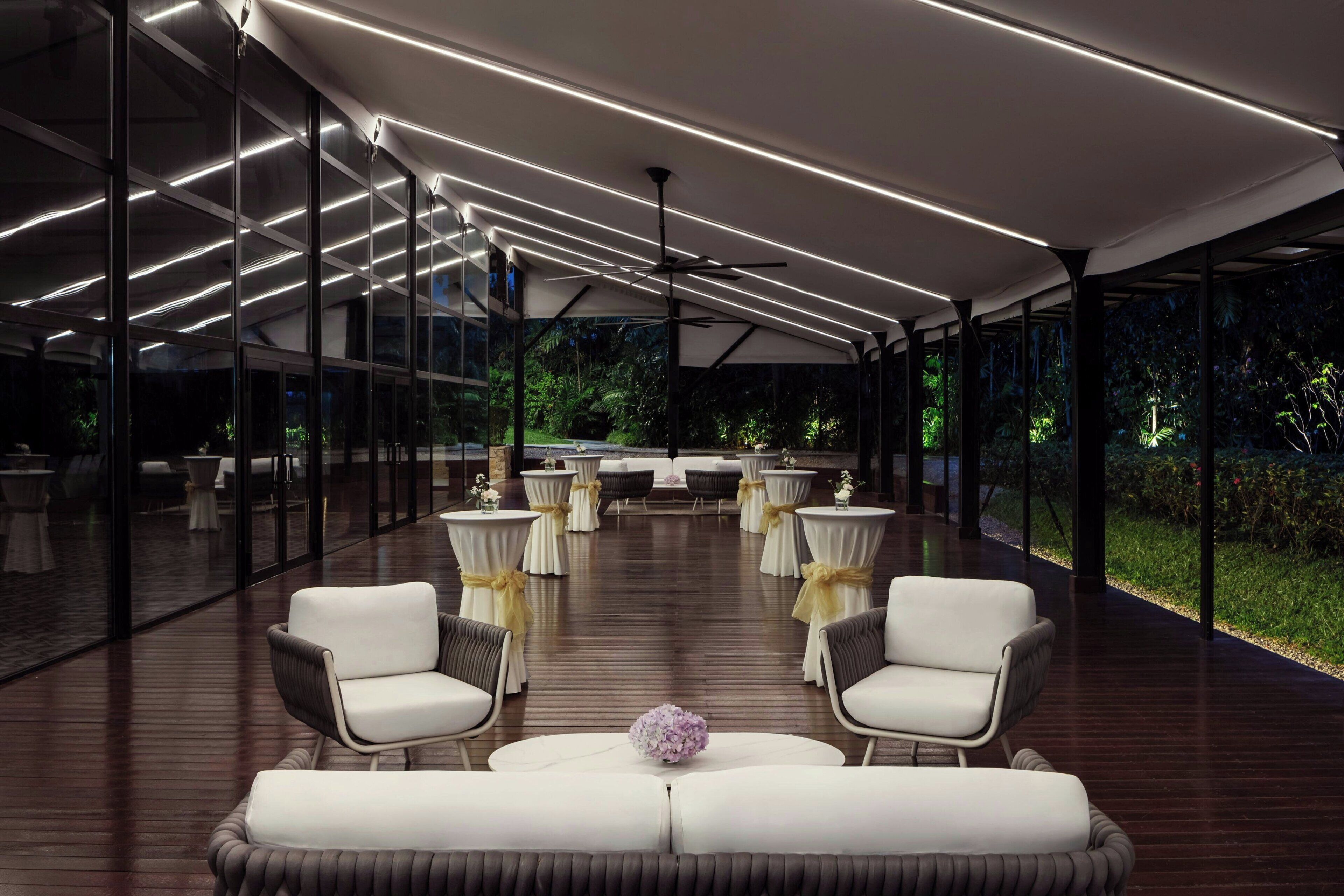 Un área lounge moderna al aire libre con paredes de vidrio, piso de madera y mobiliario blanco.