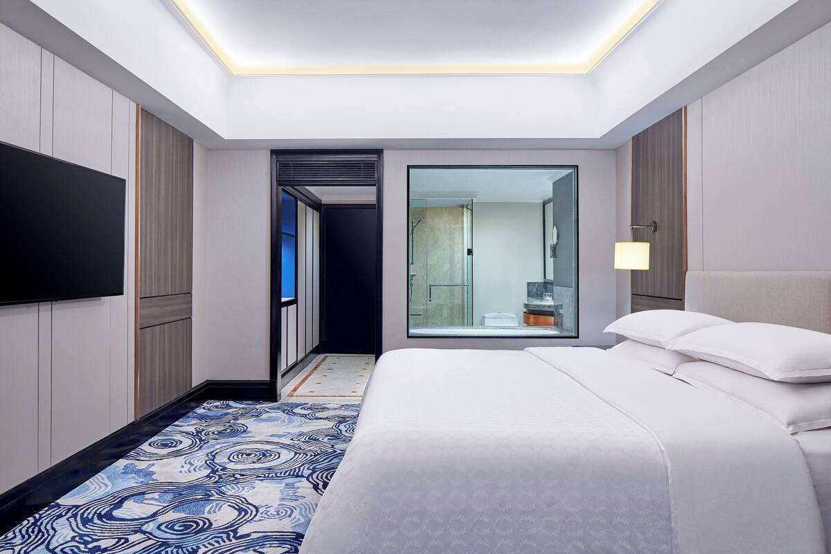 Une chambre d’hôtel moderne dotée d’un grand lit, d’une télévision à écran plat, d’un dressing et d’une salle de bain avec une douche à parois de verre.