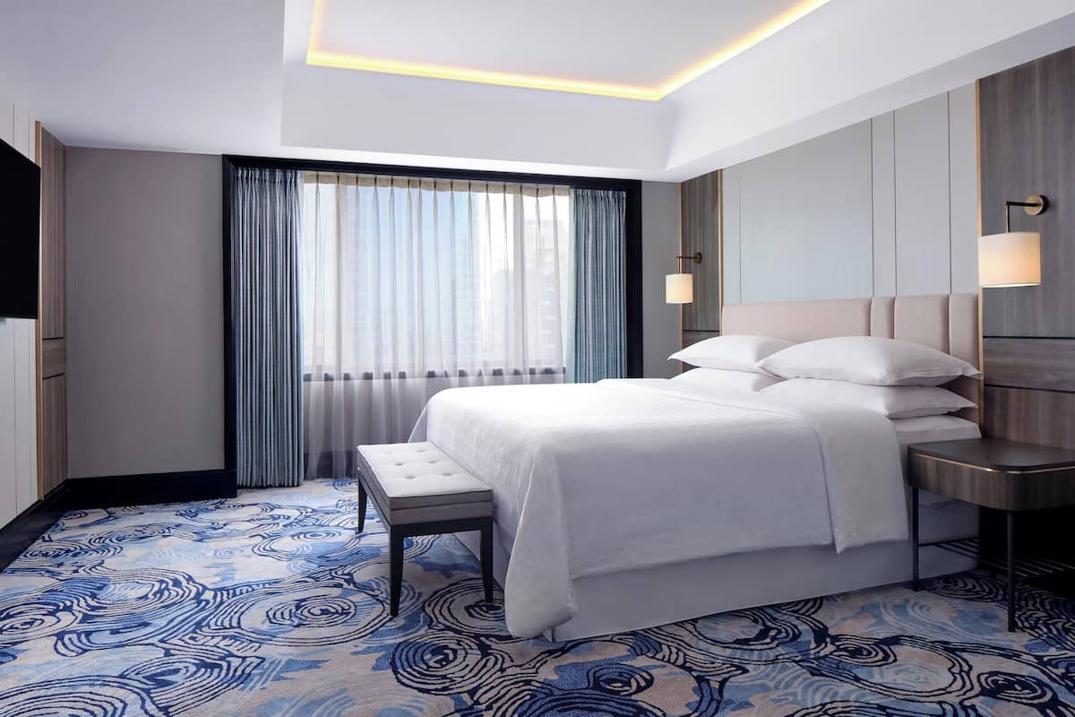 Une chambre d’hôtel moderne avec un grand lit, une banquette, une table de chevet et une grande fenêtre avec des rideaux.