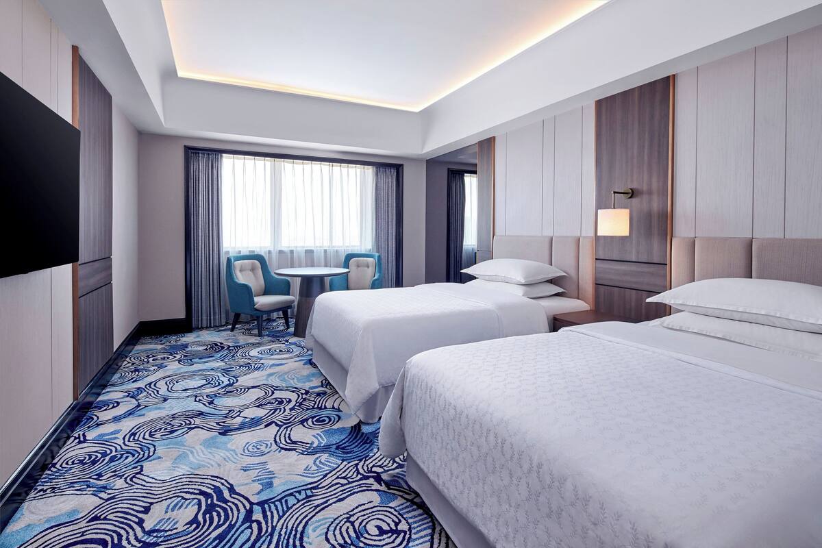 Une chambre d’hôtel avec deux lits, un téléviseur à écran plat, une petite table ronde avec une chaise, et une grande fenêtre avec des rideaux.