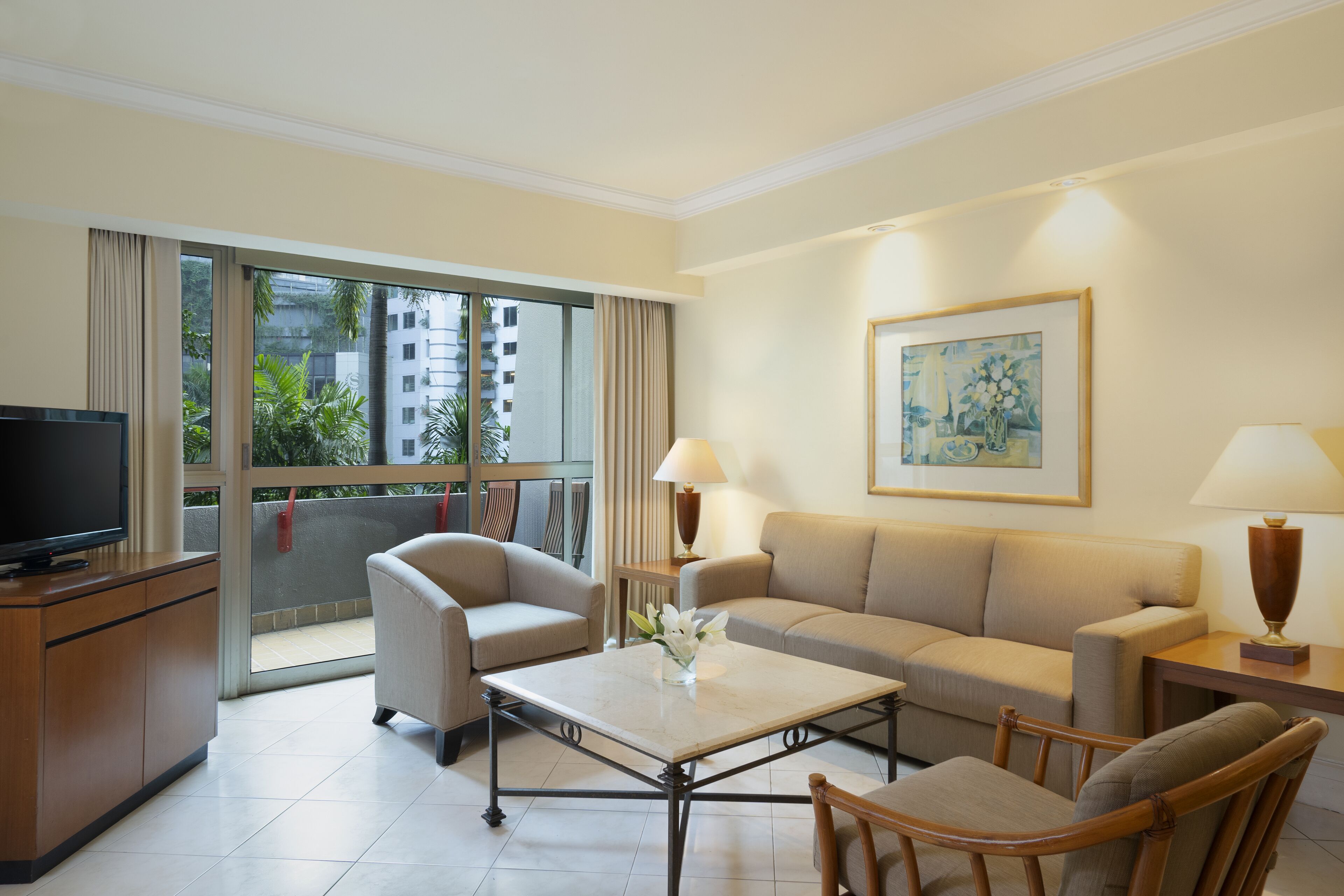 A hotel room with a sofa, armchairs, a coffee table, a TV, and a balcony with a view of buildings and greenery.