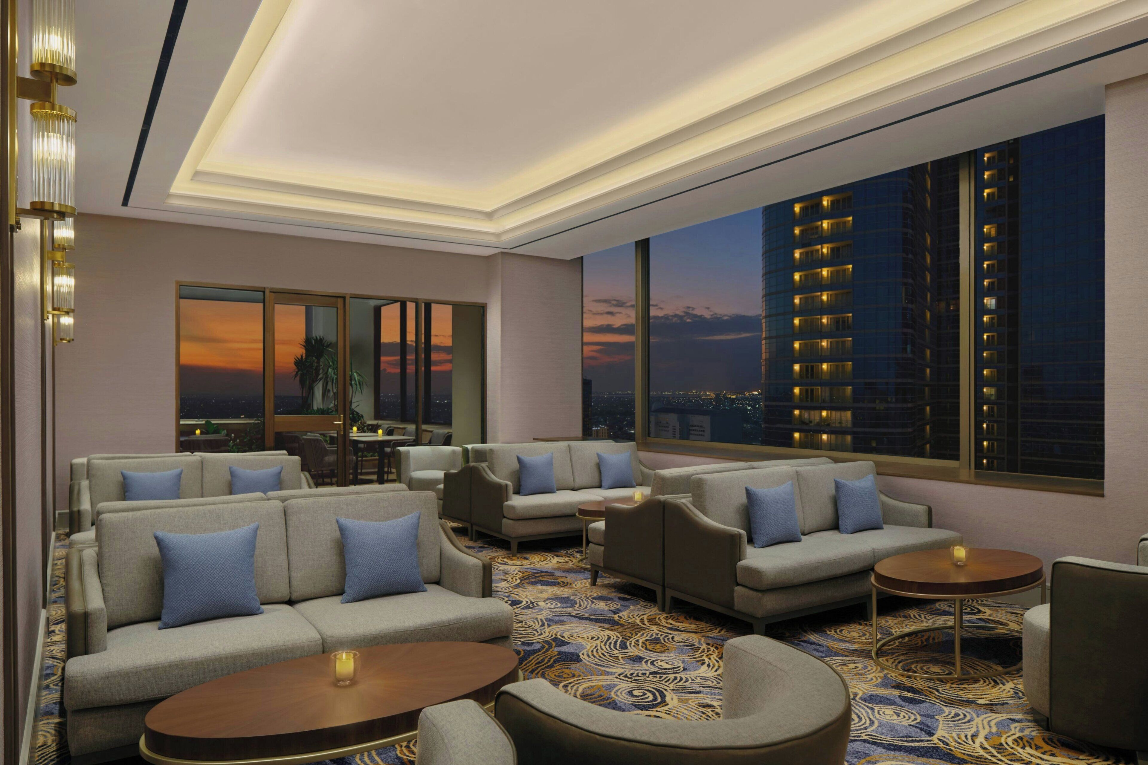 A modern lounge area with sofas, armchairs, and round tables, featuring large windows with a cityscape view.