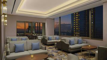A modern lounge area with sofas, armchairs, and round tables, featuring large windows with a cityscape view.