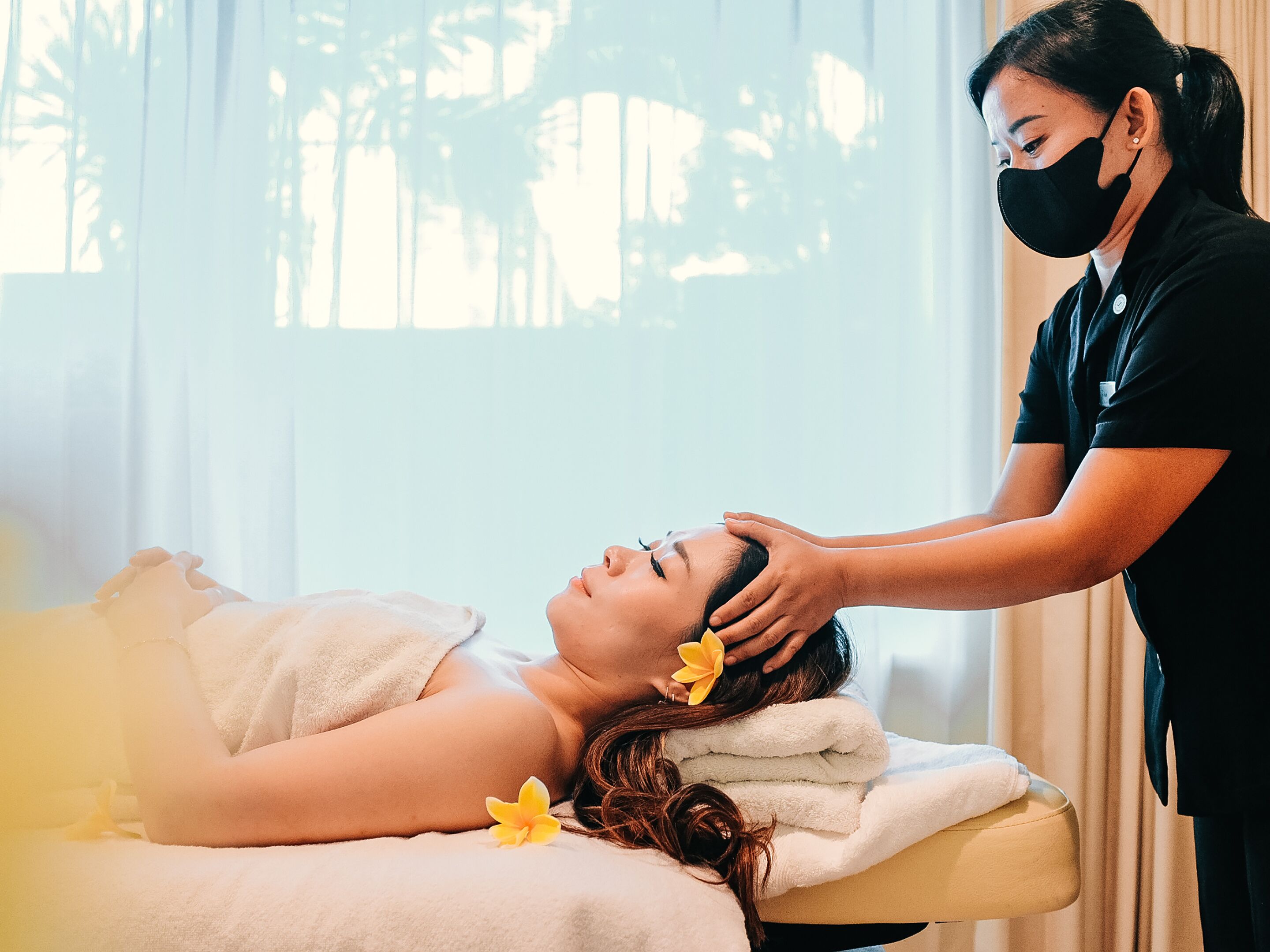 Iemand ontvangt een hoofdmassage in een kamer met raam en gordijnen.
