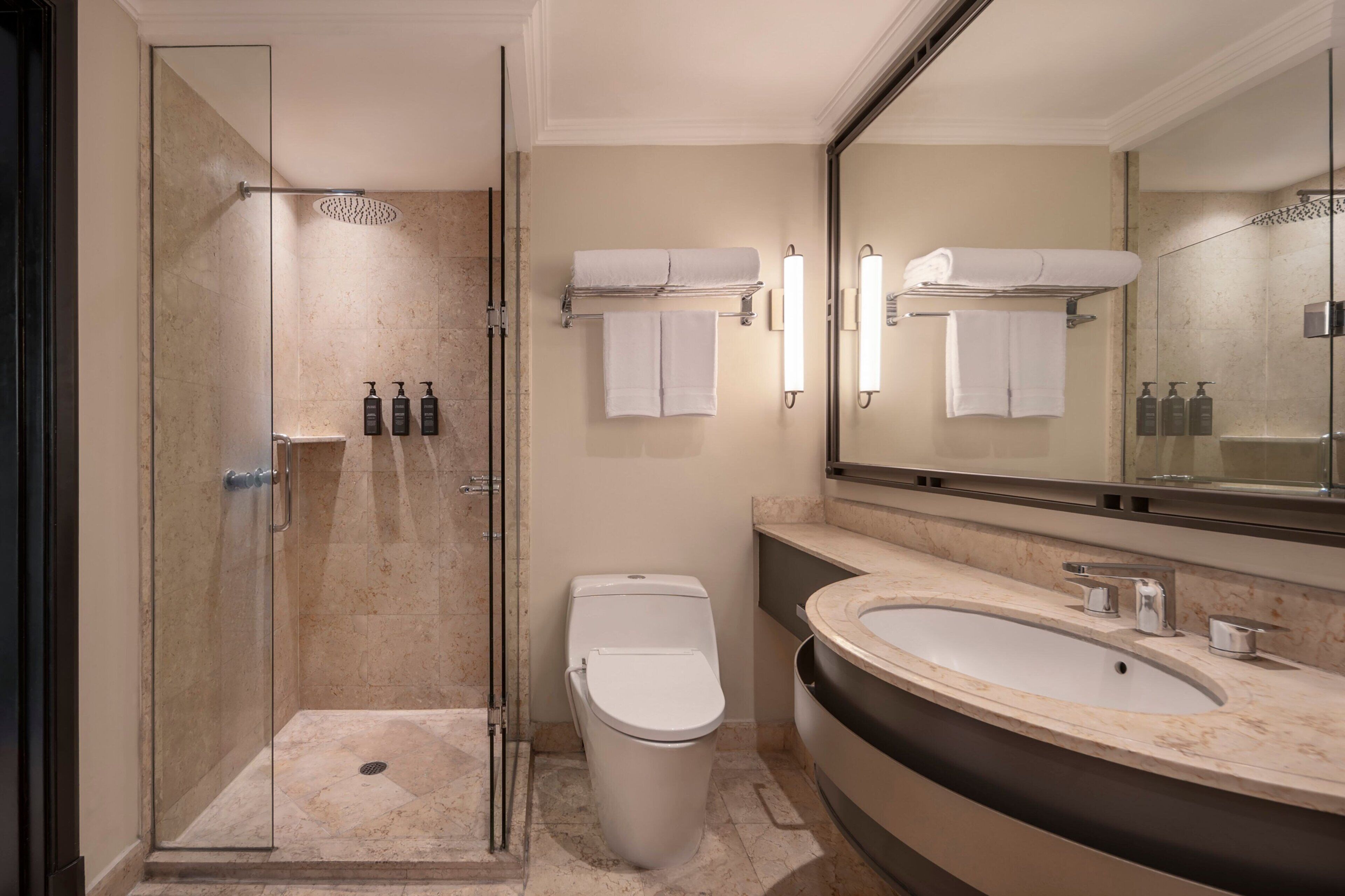 Un baño moderno con ducha envuelta en vidrio, inodoro, lavamanos con espejo y dos toalleros con toallas dobladas.