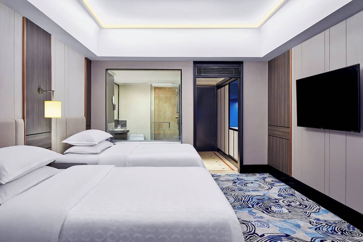 Une chambre d’hôtel avec deux lits, un téléviseur à écran plat, un miroir et une salle de bain visible à travers une porte ouverte.