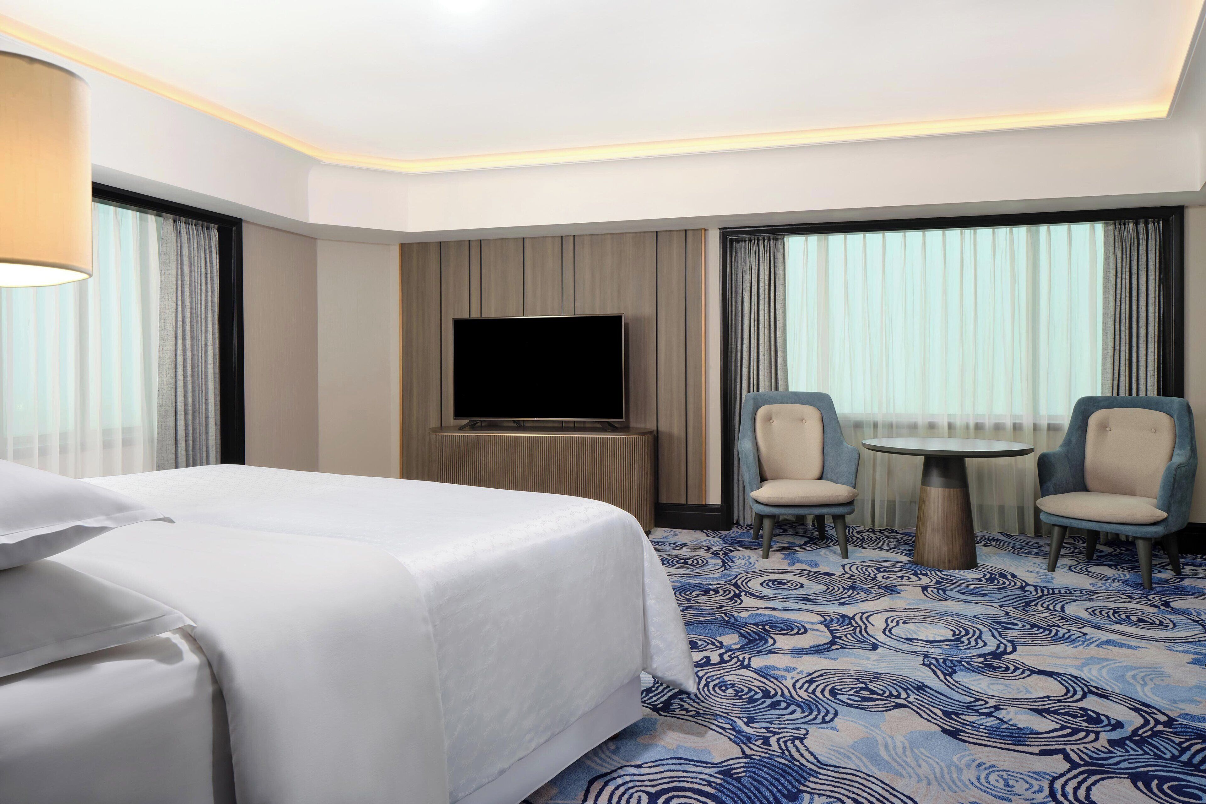 Habitación de hotel con una cama grande, un televisor de pantalla plana, dos sillones, una mesita redonda y una alfombra estampada.