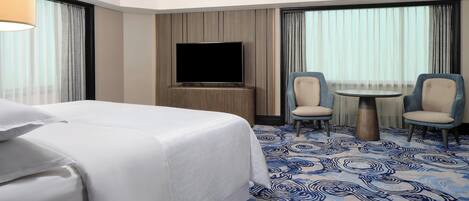 Habitación de hotel con una cama grande, un televisor de pantalla plana, dos sillones, una mesita redonda y una alfombra estampada.