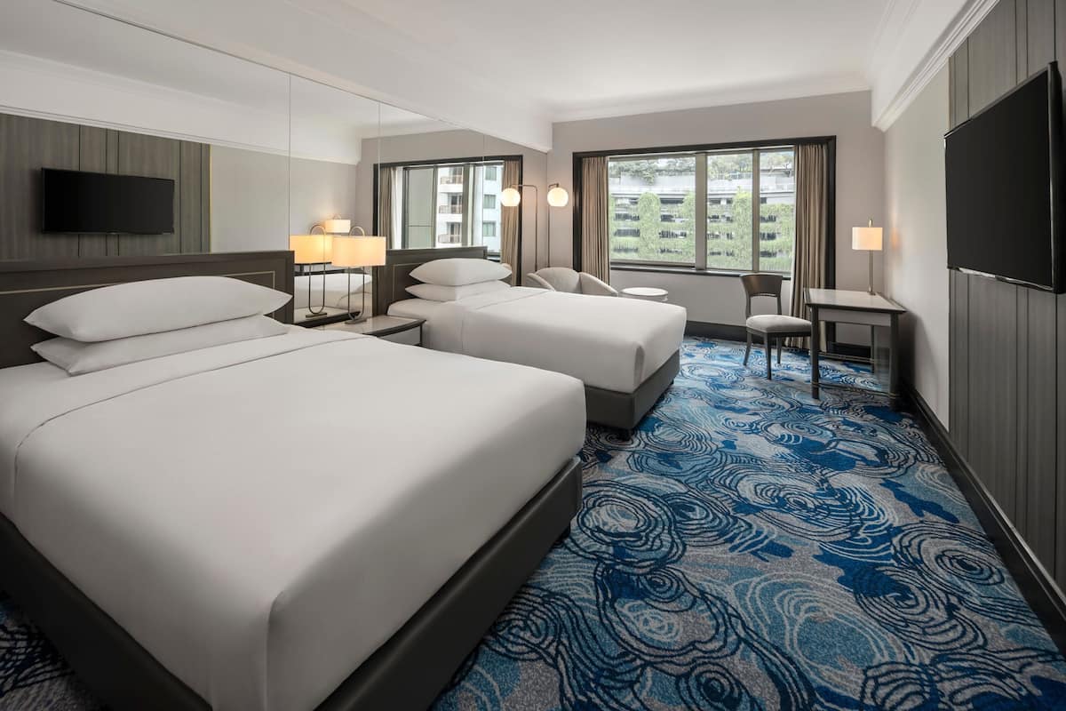 Une chambre d’hôtel avec deux lits, un bureau, une chaise, une télévision et de grandes fenêtres donnant sur des immeubles.