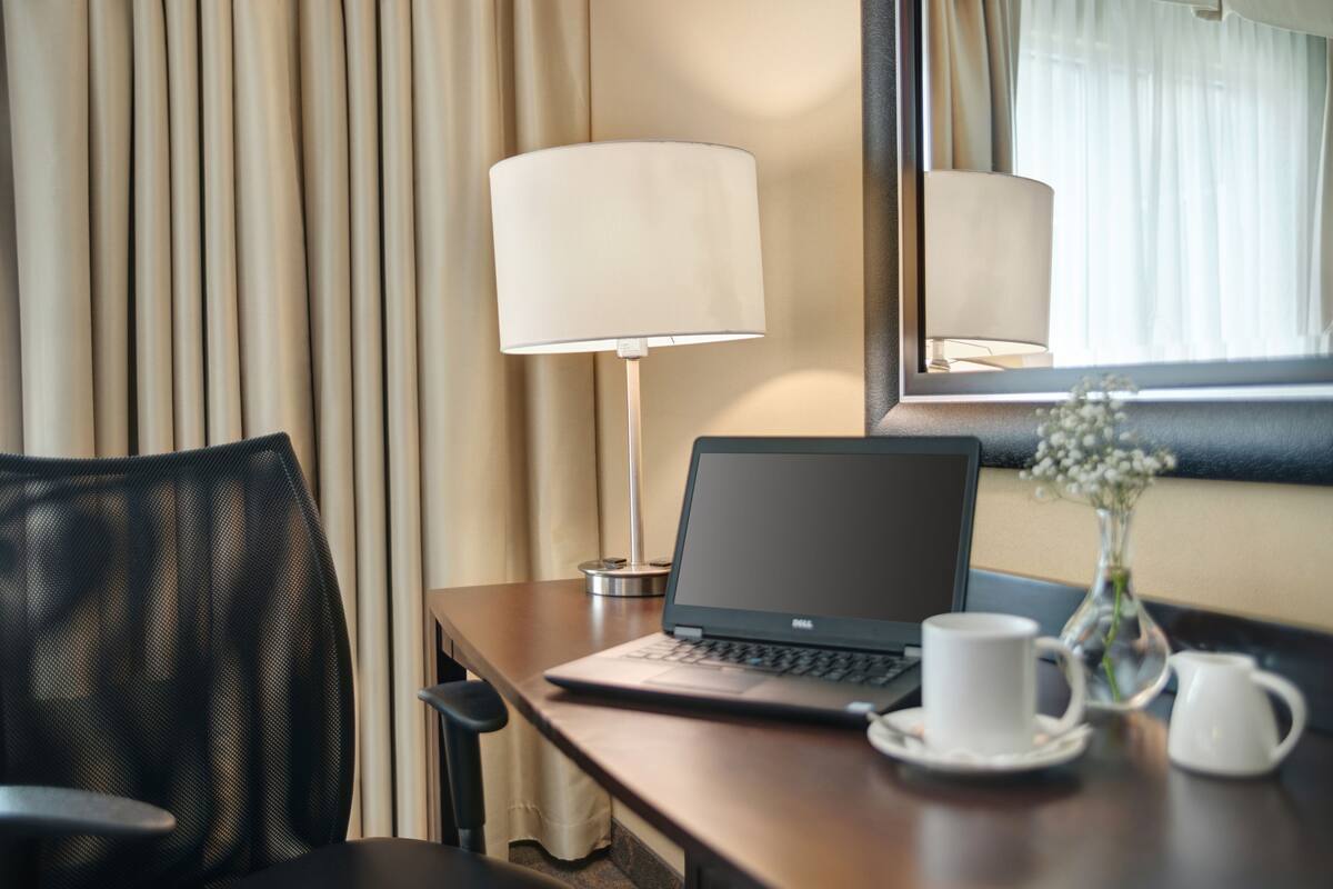 A hotel room with a desk, laptop, lamp, and a vase with flowers.