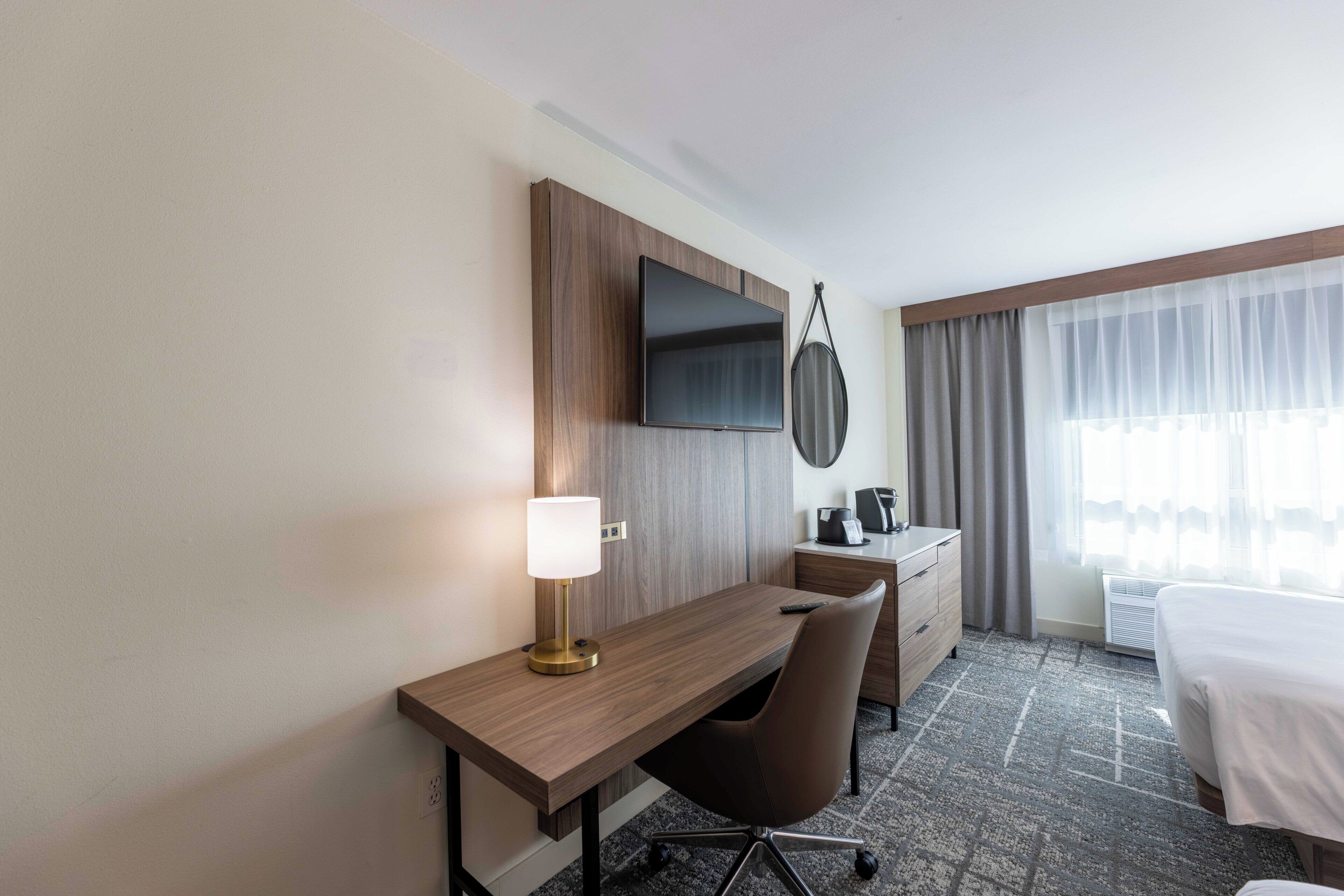 Habitación de hotel con escritorio de madera, televisor de pantalla plana en la pared, lámpara de escritorio, silla, cama, cómoda y ventana con cortinas traslúcidas.