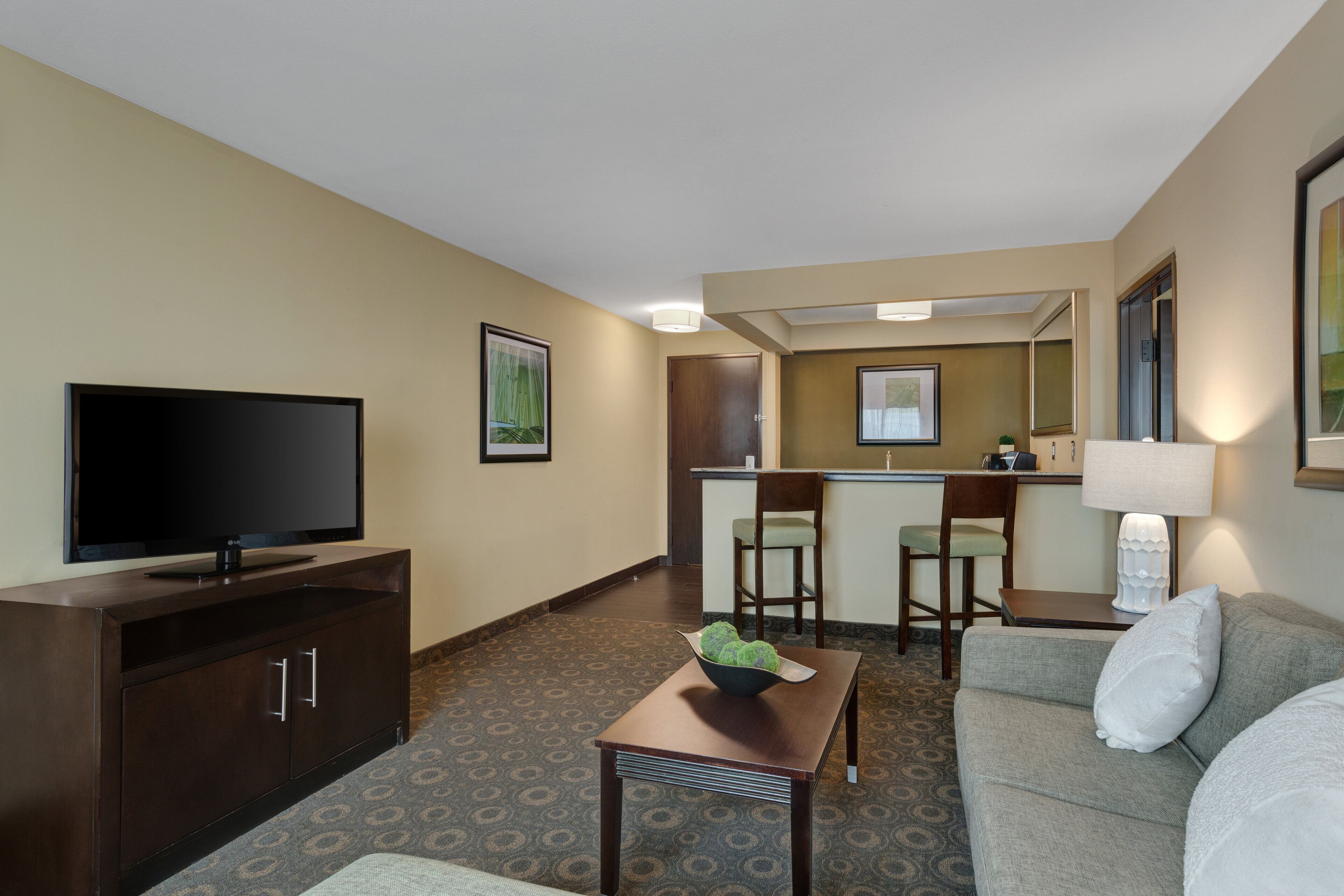 A hotel room with a flat-screen TV, a sofa, a small table with a bowl of fruit, a kitchenette, and a dining area with chairs.