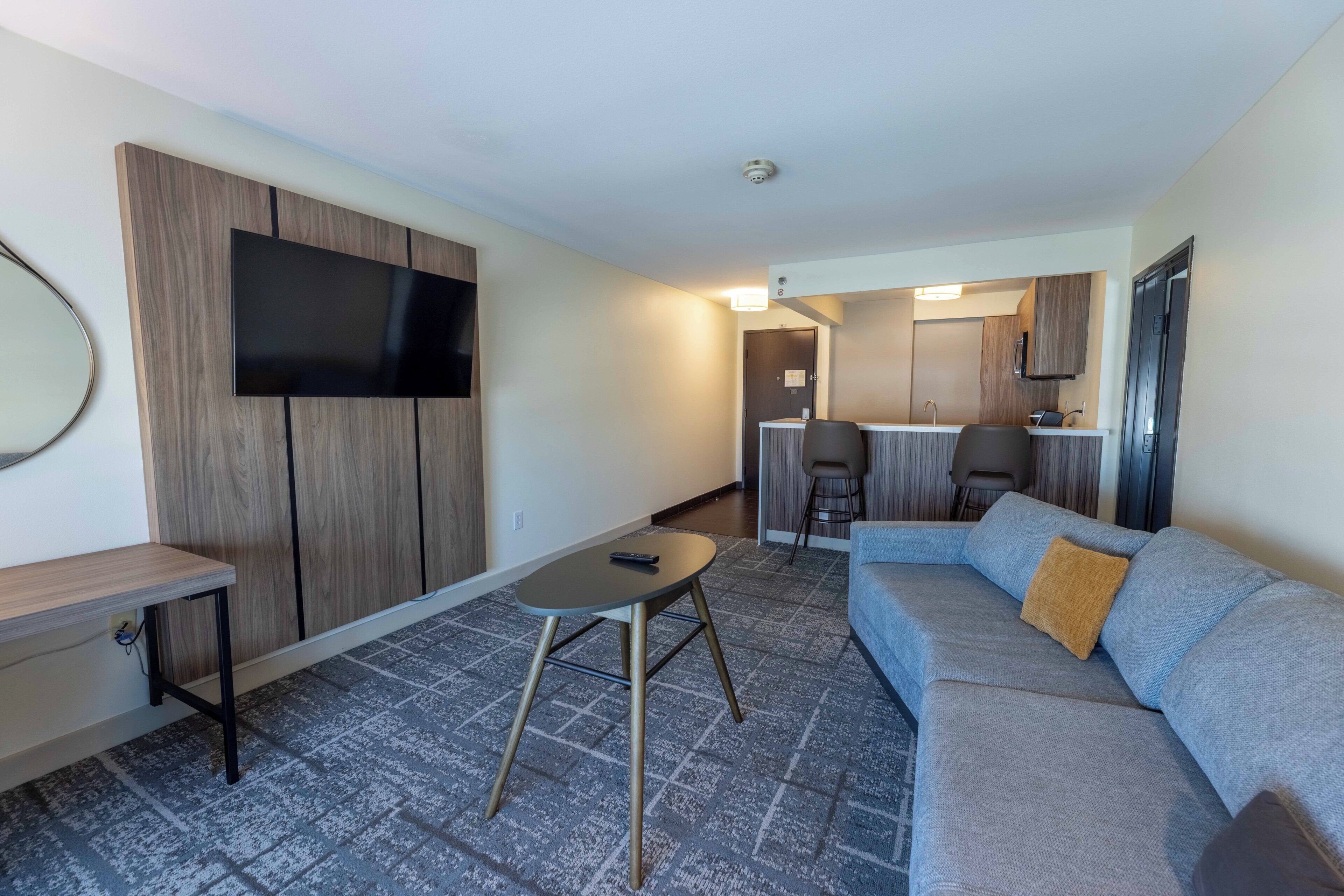 A modern hotel room with a flat-screen TV, a sofa, a small table, and a kitchenette in the background.