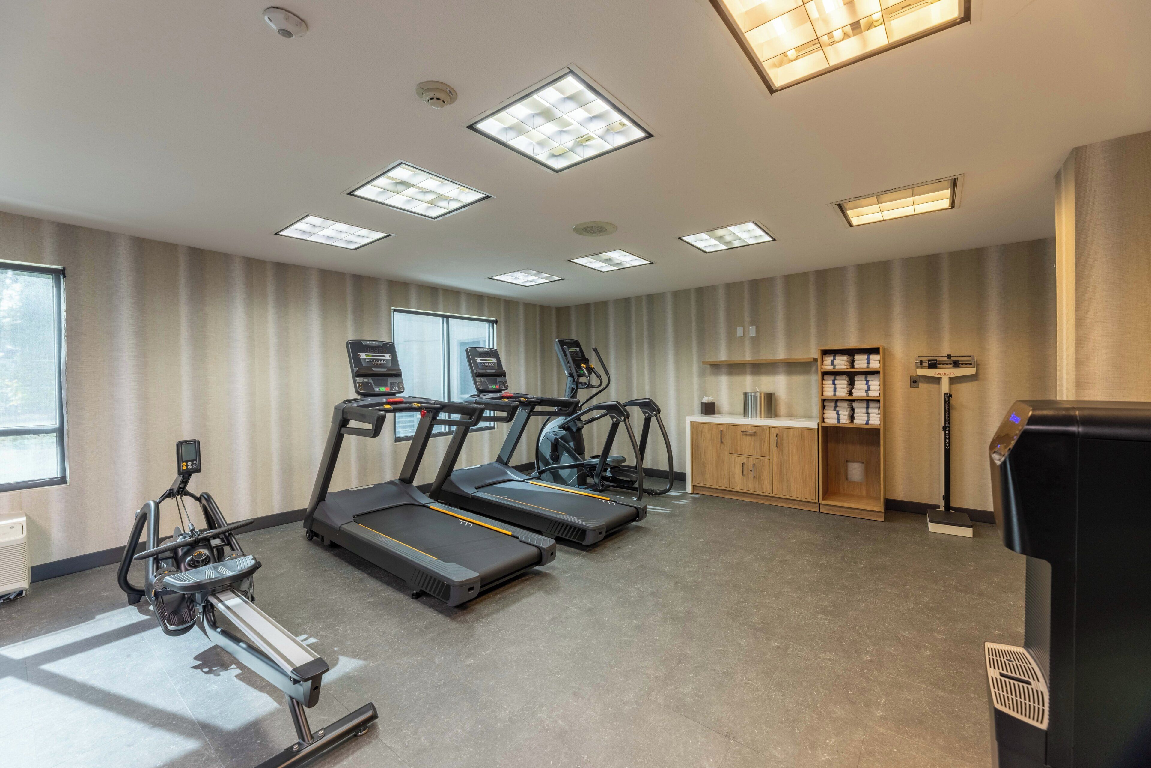 Un gimnasio con cintas de correr, una bicicleta estática y una máquina de remo. Hay luces en el techo, una ventana y un mostrador con una cafetera.