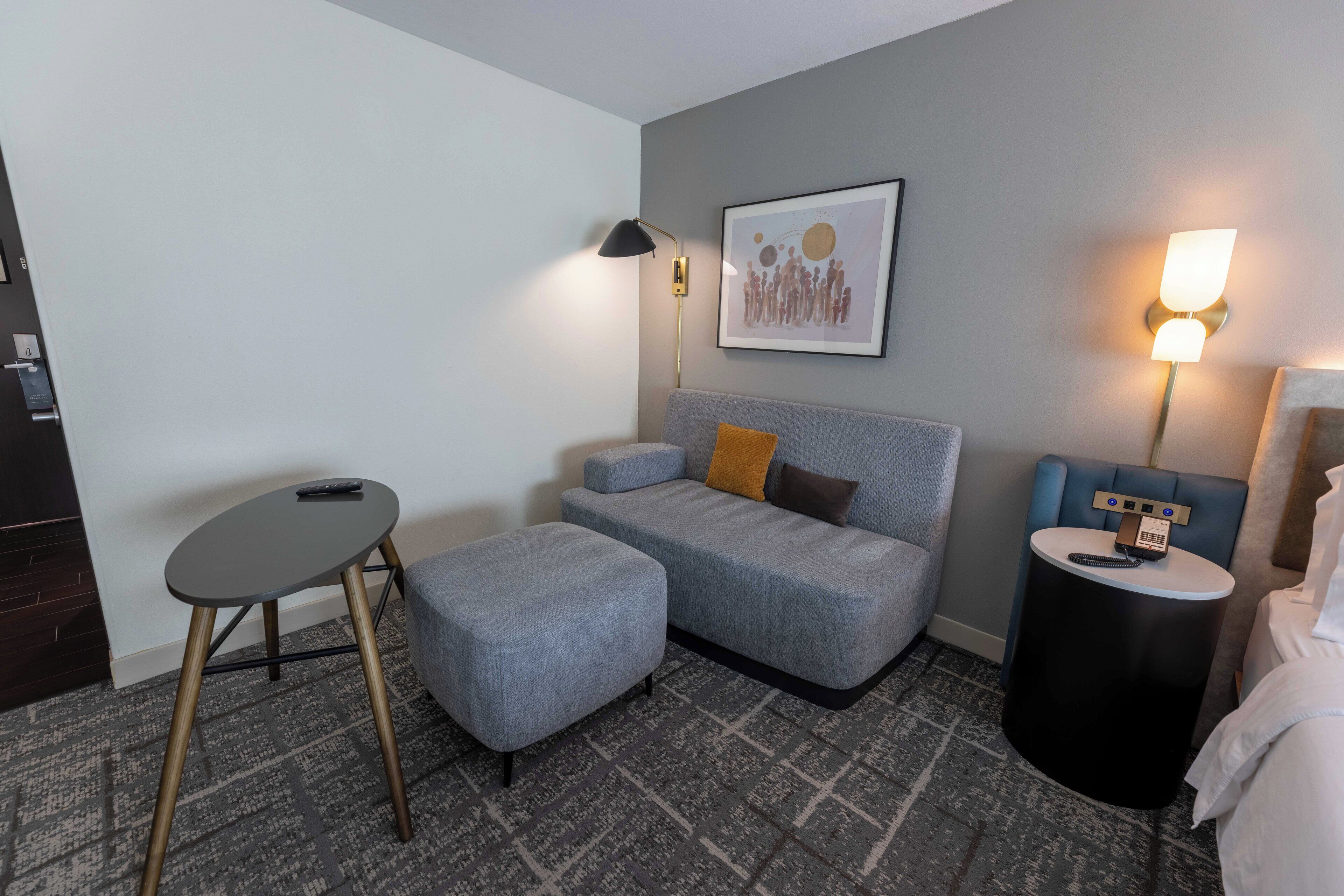 A modern hotel room with a grey sofa, a small round table, a standing lamp, and a framed artwork on the wall.