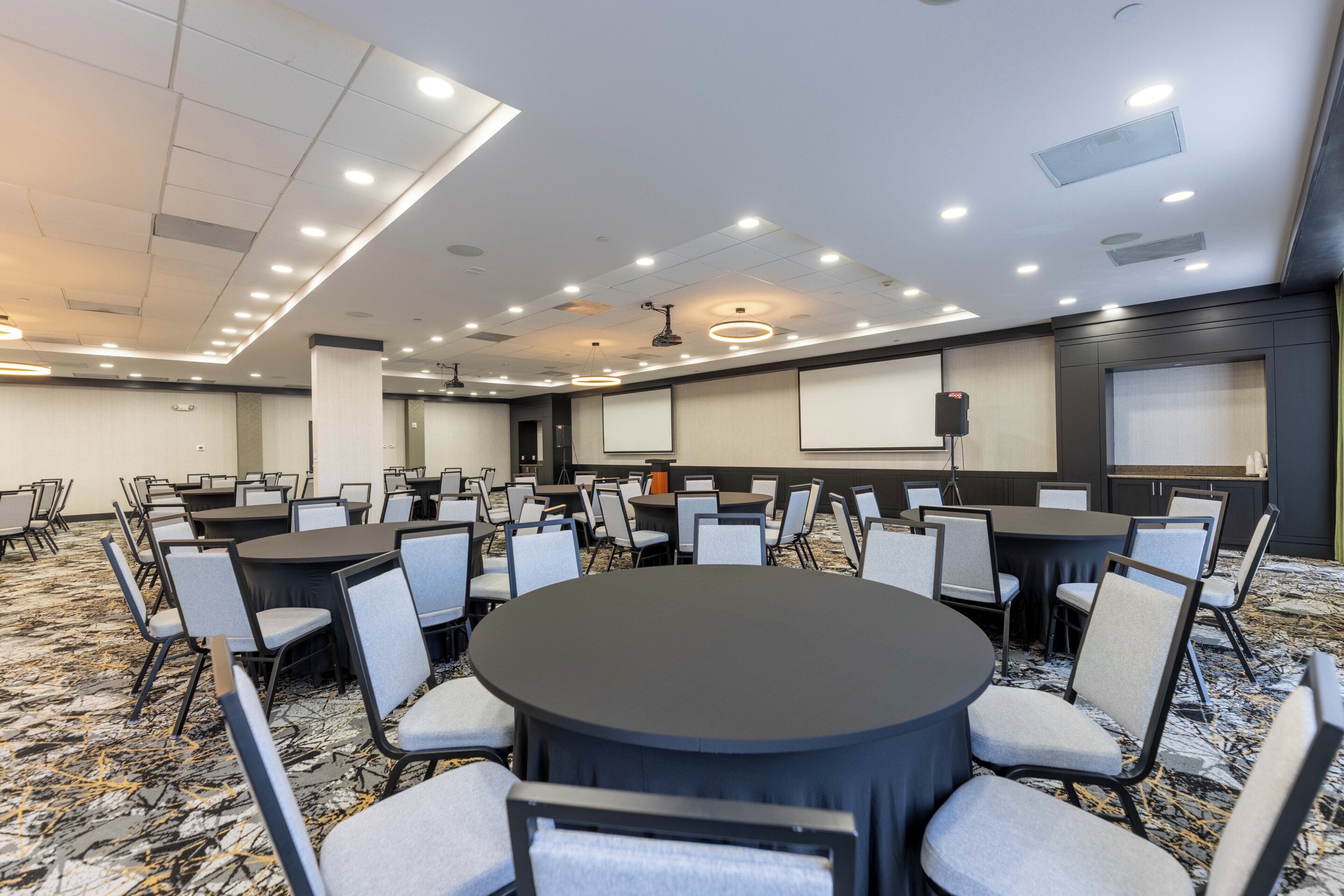 A conference room with multiple tables and chairs, ceiling lights, and a projector screen.