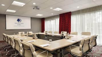 Ein Konferenzraum mit langem Tisch, beigefarbenen Stühlen, einer Projektorleinwand und einem großen Fenster mit roten Vorhängen.