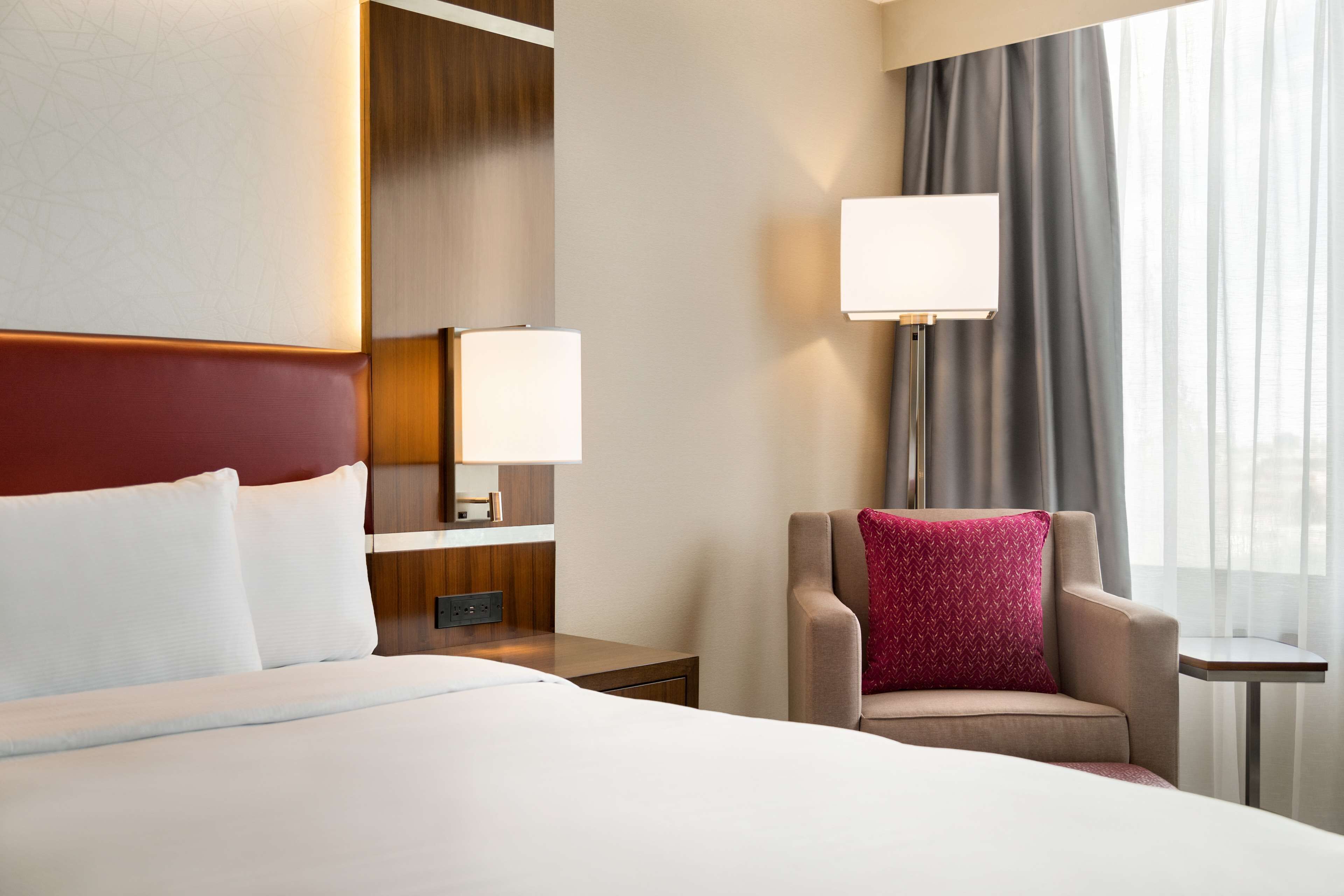 A hotel room with a large bed, a chair with a red pillow, a nightstand with a lamp, and a view of the city through the window.