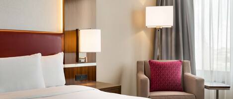 A hotel room with a large bed, a chair with a red pillow, a nightstand with a lamp, and a view of the city through the window.