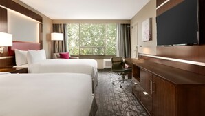 Habitación de hotel con dos camas, un escritorio, un televisor y un ventanal con vistas a árboles.