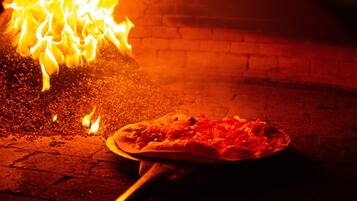 Un horno de pizza con llamas, una pizza sobre una pala y un muro de ladrillo al fondo.